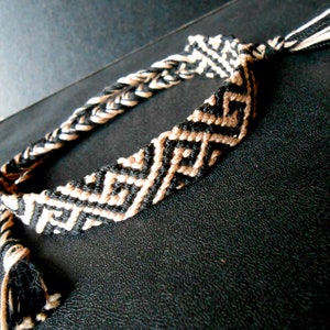 Può includere: Braccialetto dell'amicizia fatto a mano in nero e beige. Il braccialetto presenta un motivo geometrico al centro e un design intrecciato sui lati. Le estremità sono rifinite con nappe. Un accessorio elegante per l'uso quotidiano.