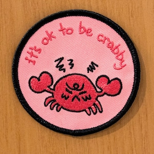Op de afbeelding: Een ronde, roze geborduurde patch met de tekst "It's ok to be crabby" in rode letters. De patch toont een cartoonkrab met rode scharen en een chagrijnige uitdrukking, omlijnd in zwart.