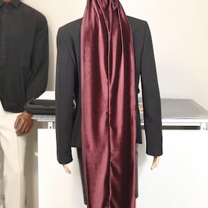 May include: A burgundy velvet durag is draped over a mannequin wearing a black blazer. The durag is long and flowing, with a tied knot at the top. The mannequin is standing in front of a white background.