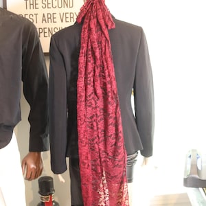 May include: A mannequin in a black blazer and skirt, adorned with a burgundy lace head covering and scarf. The scarf cascades down the back. A sign with text is visible in the background.