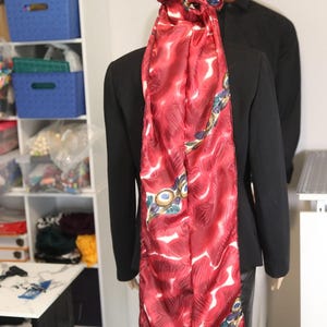 May include: A red patterned scarf draped over a black blazer. The scarf features a repeating design with white and blue accents. The scarf is tied on top of the head. A mannequin is wearing the blazer.