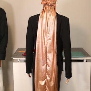 May include: A mannequin wearing a black blazer and a metallic peach-colored head covering and scarf. The head covering is tied at the back of the head, and the scarf flows down the back of the blazer.