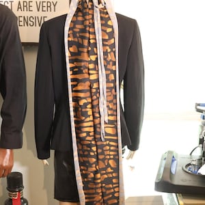 May include: A mannequin is dressed in a black blazer and skirt. A patterned head covering and scarf in black, brown, and gold tones are draped over the mannequin's head and back. The scarf has a light grey border.