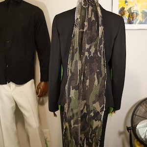 May include: A long, rectangular head covering made of a camouflage-patterned mesh fabric in shades of green, brown, and black. The fabric features a grid-like texture and is tied at the back of the head, with the long tail draping down the back.