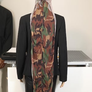 May include: A mannequin wearing a black blazer and a camouflage-patterned head covering and draping. The head covering features a mix of brown, green, and tan colours. The blazer is black, and the mannequin is on a white surface.