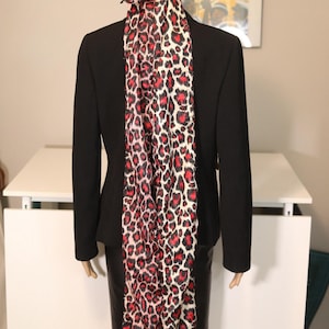 May include: A black blazer and skirt are worn with a matching leopard print head wrap and scarf. The scarf has a red and black spotted pattern on a white background. The mannequin is positioned in front of a white table.