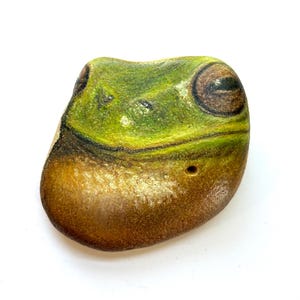 May include: A painted stone shaped like a frog's head. The top is green with black accents, transitioning to a brown base. The frog's eye is brown. A small hole is visible near the bottom.