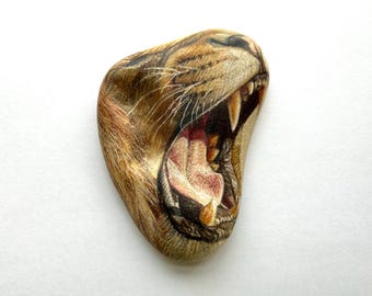 May include: Close-up of a painted rock depicting a lion's face with its mouth open, showing its teeth. The rock is painted in shades of brown, yellow, and red.