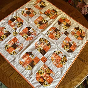 May include: A square quilted table topper featuring a grid of nine octagonal blocks separated by white fabric strips. The blocks display a seasonal pattern of orange pumpkins, sunflowers, and plaid fabric in shades of brown, orange, and yellow.