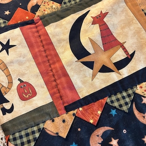 Könnte beinhalten: Ein gesteppter Stoffläufer mit Patchwork-Design und Halloween-Motiven, darunter orangefarbene Mondsicheln, schwarze Sterne, eine rot gestreifte Katze auf einem goldenen Stern und ein kleiner roter Kürbis mit schwarz-grünen Karomustern.