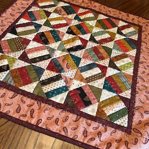 Könnte beinhalten: Ein quadratischer, gesteppter Tischläufer mit einem Patchwork-Design aus mehrfarbigen Stoffstreifen in Rot-, Grün-, Gold- und Cremetönen. Die Mitte ist von einem breiten Rand aus pfirsichfarbenem Stoff mit dunkelbraunem Paisleymuster umrahmt.