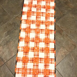 May include: A rectangular table runner featuring a patchwork design of alternating white squares and orange plaid squares. The orange plaid fabric displays a repeating pattern of small pumpkins. The runner is finished with a dark brown border.