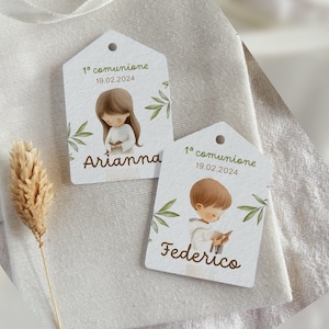 Può includere: Due etichette regalo bianche con illustrazioni di bambini in abito da comunione. Le etichette presentano il testo "1a comunione 19.02.2024" e i nomi "Arianna" e "Federico" in corsivo, con accenti di foglie verdi.