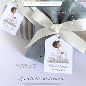 Op de afbeelding: Close-up van verschillende kleine stoffen zakjes vastgebonden met witte linten. Elk zakje heeft een gepersonaliseerd label met een kinderillustratie en de tekst "Prima Comunione Riccardo 07.06.2025". De zakjes zijn in verschillende kleuren.