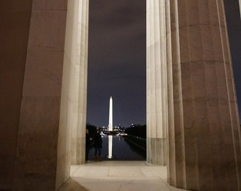 May include: A view of the Washington Monument through the columns of the Lincoln Memorial. The monument is illuminated at night, and the reflection of the lights can be seen in the water.