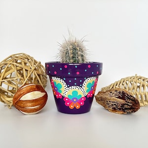 Könnte beinhalten: Lilafarbener Terrakotta-Blumentopf mit einem weißen Kaktus und einem farbenfrohen Blumendesign. Der Topf hat ein weißes Punkte-Muster und einen gewellten Rand.