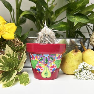 Peut inclure: Un petit cactus dans un pot peint en rouge avec un motif de poires colorées. Le pot est entouré de plantes vertes, de fleurs jaunes et de poires. Le cactus a des épines blanches.