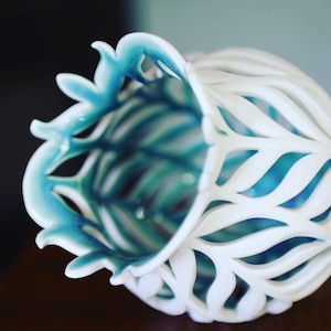 Puede incluir: Un jarrón de cerámica blanca con un esmalte azul, que presenta un delicado y complejo diseño de hojas superpuestas. El jarrón está abierto en la parte superior y tiene una base ancha y redondeada.