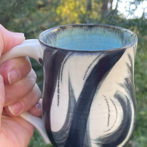Può includere: Una tazza in ceramica bianca con un design marmorizzato bianco e nero. La tazza ha una glassa blu chiaro all'interno e un bordo nero.