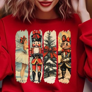 May include: A red sweatshirt featuring four vertical panels with Christmas-themed illustrations. The panels depict a ballerina, a nutcracker, a Christmas tree, and a mouse in a military-style outfit. The sweatshirt has long sleeves.
