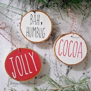 May include: Three round wooden ornaments with red and white paint. The ornaments are decorated with the words "Bah, Humbug", "Jolly", and "Cocoa".