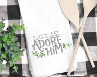 Pode incluir: Uma toalha de cozinha branca com o texto "O come let us adore him" em letras cinzas com folhas verdes de cada lado do texto. A toalha está dobrada e colocada em uma cesta de madeira com um pano xadrez preto e branco por baixo. Duas colheres de madeira são colocadas em cima da toalha.