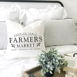 May include: White pillow with black text that reads "Farm to Table Always Fresh Farmers Market Dairy Produce Dry Goods". The pillow is on a white bed with a gray and white striped pillow behind it. A small potted plant is in the foreground.