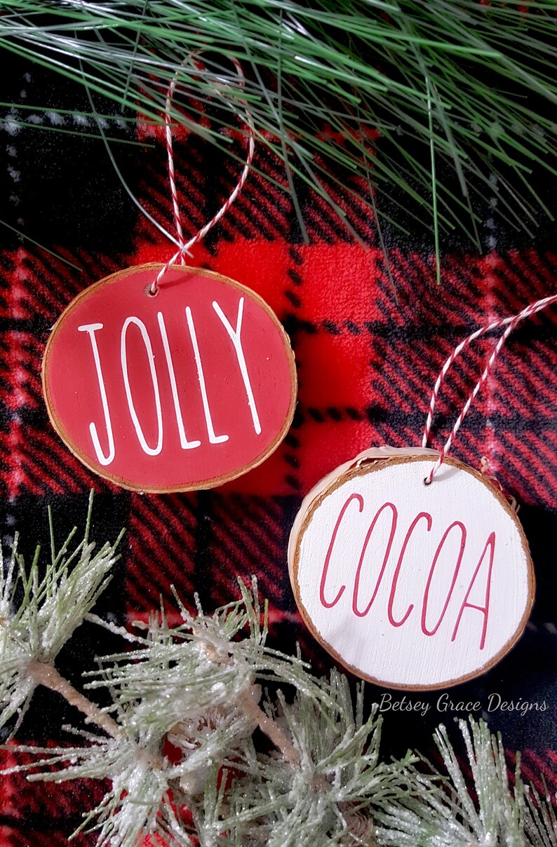 May include: Two round wooden ornaments with red and white paint. The red ornament says "Jolly" in white paint. The white ornament says "Cocoa" in red paint. The ornaments are on a red and black plaid fabric background.