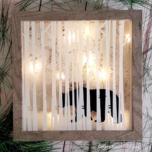 May include: A wooden frame with a white silhouette of trees and a black bear inside. The frame has a light brown finish and is illuminated by warm white LED lights.