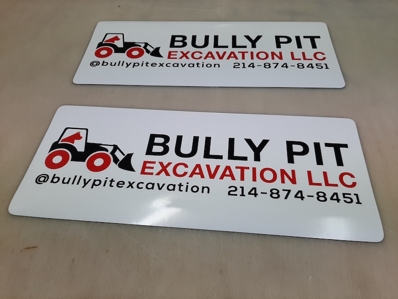 May include: White sign with black and red text and a black and red illustration of a bulldozer. The text reads "Bully Pit Excavation LLC" and includes the phone number "214-874-8451" and the social media handle "@bullypitexcavation".
