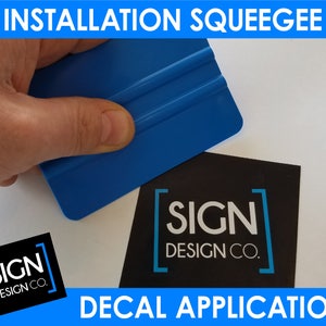 May include: A blue squeegee being used to apply a decal to a black surface with the text "SIGN DESIGN CO." printed on it.
