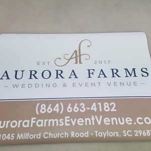 May include: White sign with brown text and a black logo. The text reads "EST 2017 AURORA FARMS - WEDDING & EVENT VENUE - (864) 663-4182 AuroraFarmsEventVenue.com 1045 Milford Church Road Taylors, SC 29687"