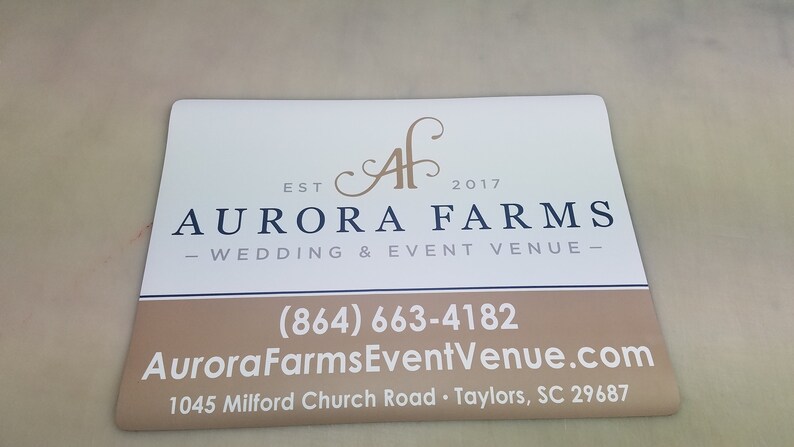 May include: White sign with brown text and a black logo. The text reads "EST 2017 AURORA FARMS - WEDDING & EVENT VENUE - (864) 663-4182 AuroraFarmsEventVenue.com 1045 Milford Church Road Taylors, SC 29687"