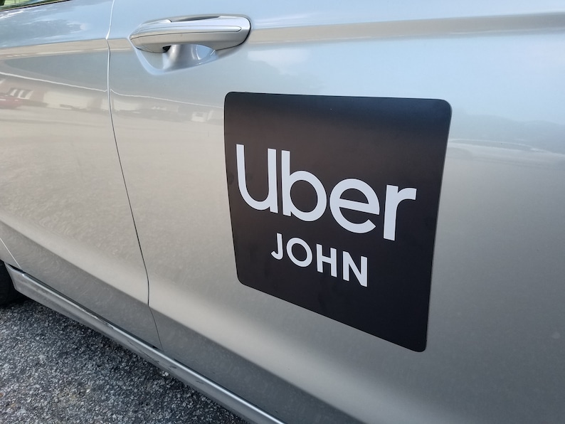 May include: A silver car with a black Uber magnet on the side door. The magnet has the Uber logo and the name "JOHN" in white text.