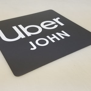 May include: A black square sign with white text that reads "Uber JOHN".