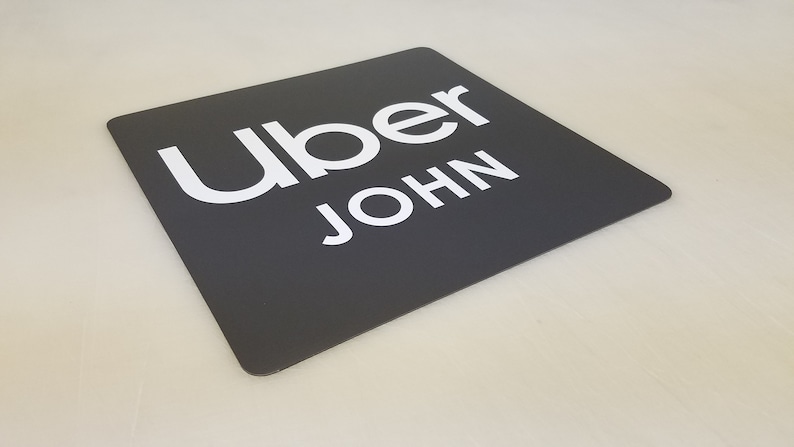 May include: A black square sign with white text that reads "Uber JOHN".