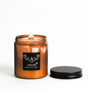 May include: A lit, orange-tinted glass jar candle with a black lid. The candle has a black label with gold text that reads "76008 CANDLE CO." The flame is bright and the jar is sitting on a white surface.