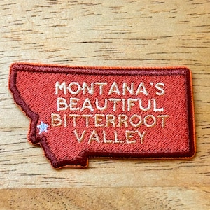 Puede incluir: Un parche rojo bordado con la forma de Montana, con el texto "MONTANA'S BEAUTIFUL BITTERROOT VALLEY" en hilo dorado. Una pequeña estrella blanca y azul está bordada en el parche. El parche está sobre una superficie de madera.