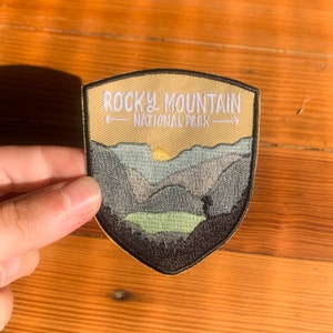 May include: A beige and black embroidered patch with the text "Rocky Mountain National Park" and a depiction of a mountain range with a sunset.