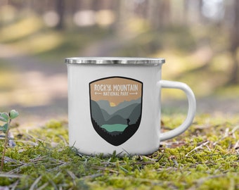 May include: White enamel camping mug with a black and brown graphic of a mountain range and a hiker. The graphic includes the text "Rocky Mountain National Park".