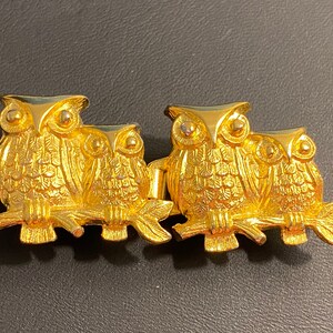 Pode incluir: Um broche de metal dourado com quatro corujas empoleiradas em um galho. As corujas são detalhadas com penas texturizadas e têm olhos pretos.