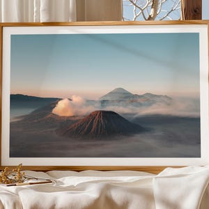Könnte beinhalten: Gerahmter Landschaftsdruck mit einer vulkanischen Bergkette. Das Bild zeigt einen Sonnenaufgang oder -untergang, mit einem Himmel, der von Blau zu Orange übergeht. Wolken und Nebel umgeben die Berge und schaffen eine ruhige Szene.