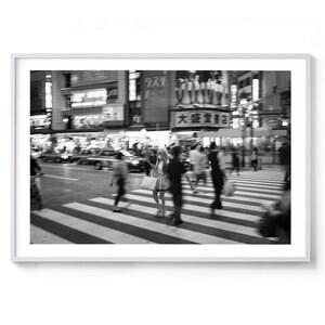 May include: Black and white photograph of a busy street scene in Japan. People are walking on a crosswalk, with blurred motion. Buildings with Japanese signage and lights are in the background. The image is framed in a white border.