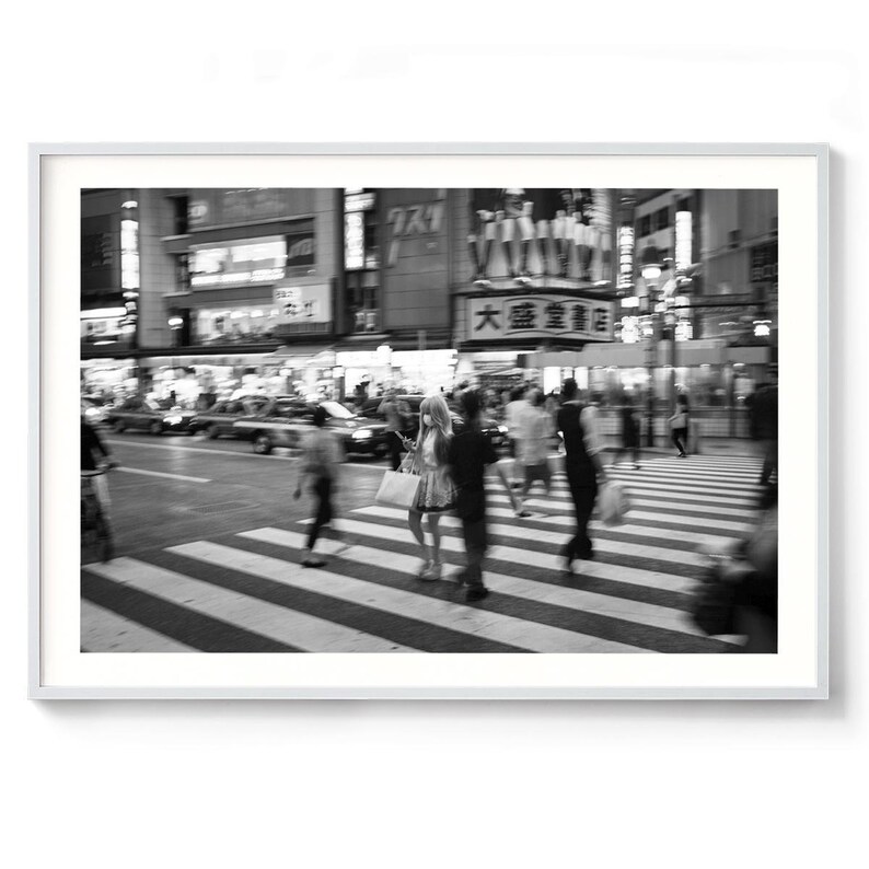 May include: Black and white photograph of a busy street scene in Japan. People are walking on a crosswalk, with blurred motion. Buildings with Japanese signage and lights are in the background. The image is framed in a white border.