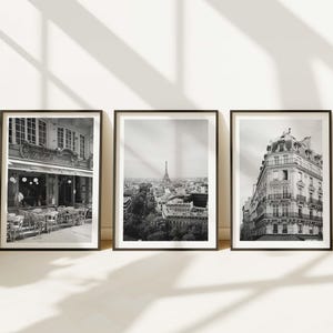Może przedstawiać: Trzy oprawione czarno-białe fotografie scen paryskich. Jedna przedstawia kawiarnię, druga wieżę Eiffla, a trzecia fasadę budynku. Ramki są czarne, a zdjęcia wyświetlane są na białej ścianie z cieniami.