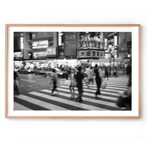 May include: Black and white photograph of a busy city street scene. People are crossing a crosswalk, with blurred motion. Buildings and signs are visible in the background, creating a sense of urban activity.