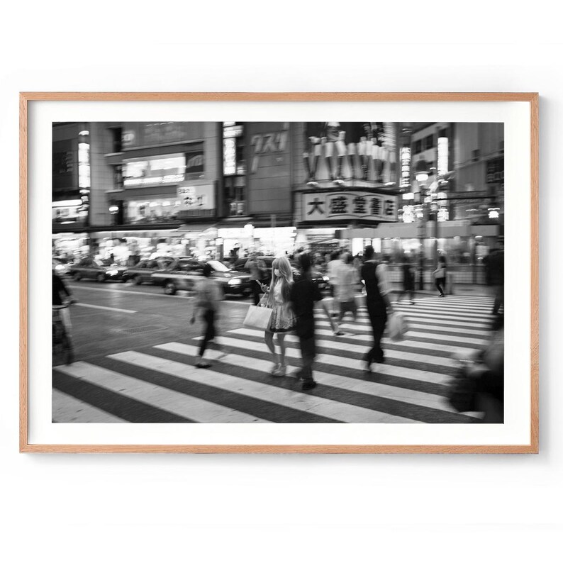 May include: Black and white photograph of a busy city street scene. People are crossing a crosswalk, with blurred motion. Buildings and signs are visible in the background, creating a sense of urban activity.