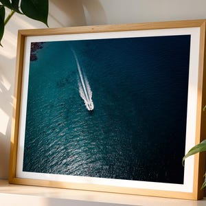 May include: A rectangular framed wall art print featuring an aerial view of a small white boat travelling across deep blue ocean water, leaving a long white wake behind it. The print is encased in a light-toned wooden frame with a white mat border.
