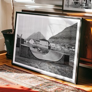May include: Black and white photograph of a half-pipe ramp in a small town. The ramp is made of concrete and is surrounded by grass. There are buildings in the background, and a mountain in the distance.