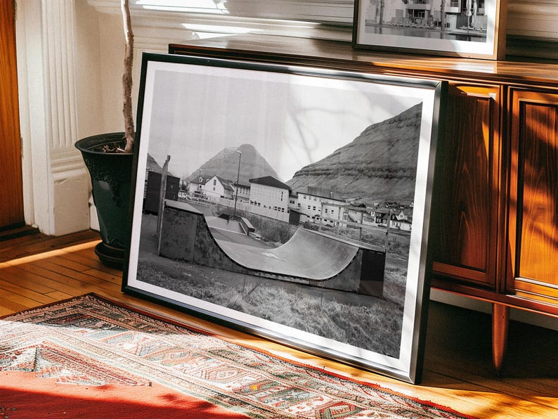 May include: Black and white photograph of a half-pipe ramp in a small town. The ramp is made of concrete and is surrounded by grass. There are buildings in the background, and a mountain in the distance.